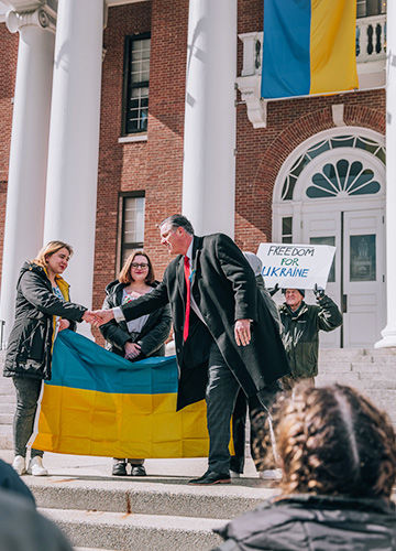 ڿ President Fred Clark shaking hands with students holding the Ukrainian flag in front of Boyden Hall; a man holds a sign that says Freedom for Ukraine